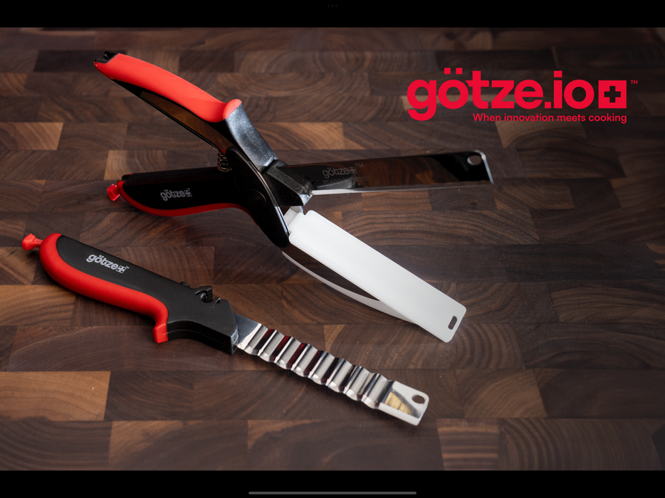 Götze multi-function kitchen shears with stainless steel blade and white safety guard displayed with a matching red and black crinkle cutter on a dark wooden cutting board.