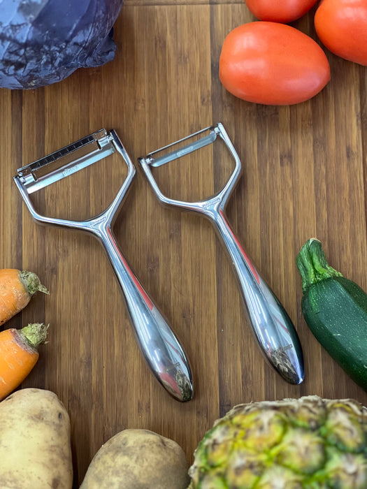 Götze Luxury Premium Peeler with polished ergonomic handles displayed on a wooden cutting board surrounded by fresh vegetables.