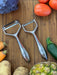 Götze Luxury Premium Peeler with polished ergonomic handles displayed on a wooden cutting board surrounded by fresh vegetables.
