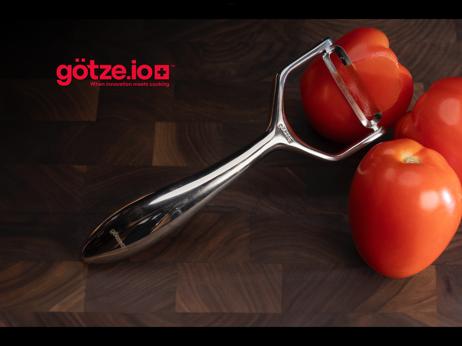 Götze Luxury Premium Peeler removing thin skin from a fresh tomato on a dark wooden cutting board.