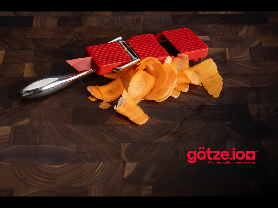 Götze Luxury Premium Peeler with freshly sliced carrot ribbons on a dark wooden cutting board.