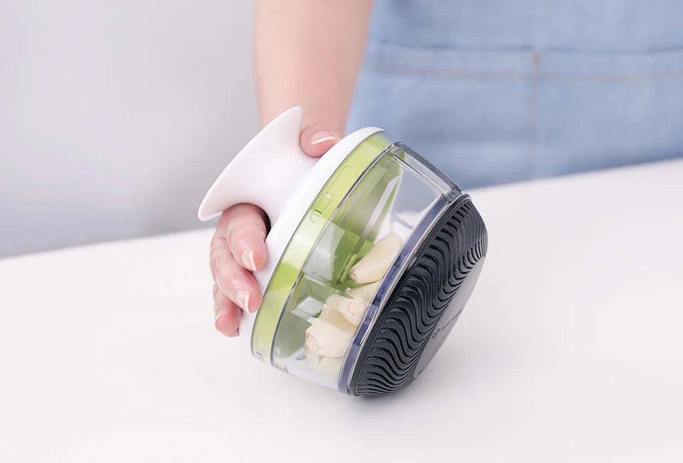 Götze Mavo manual food chopper held in hand with garlic cloves inside the clear container, featuring green blades and a black non-slip base.