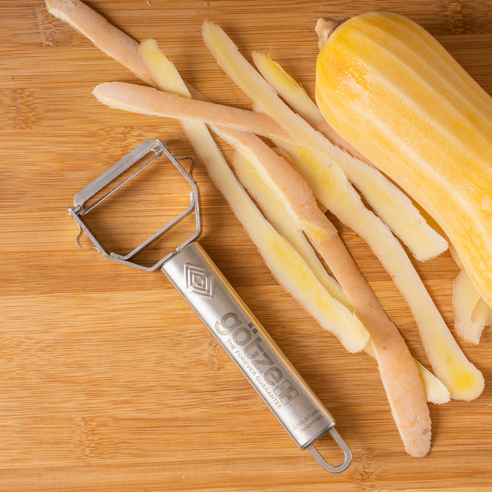 Götze stainless steel peeler shown beside peeled squash and long peel strips on a wooden cutting board.