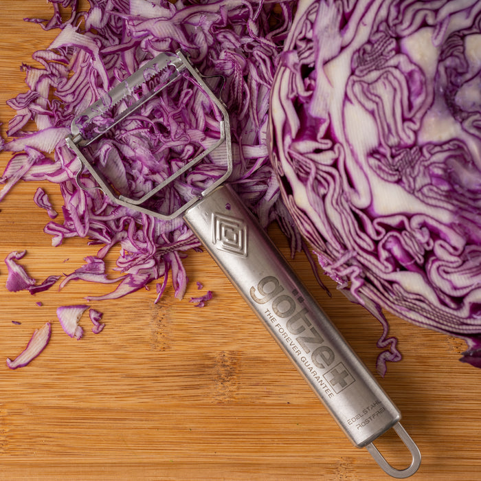 Götze stainless steel julienne peeler displayed beside shredded red cabbage and a halved cabbage on a wooden cutting board.