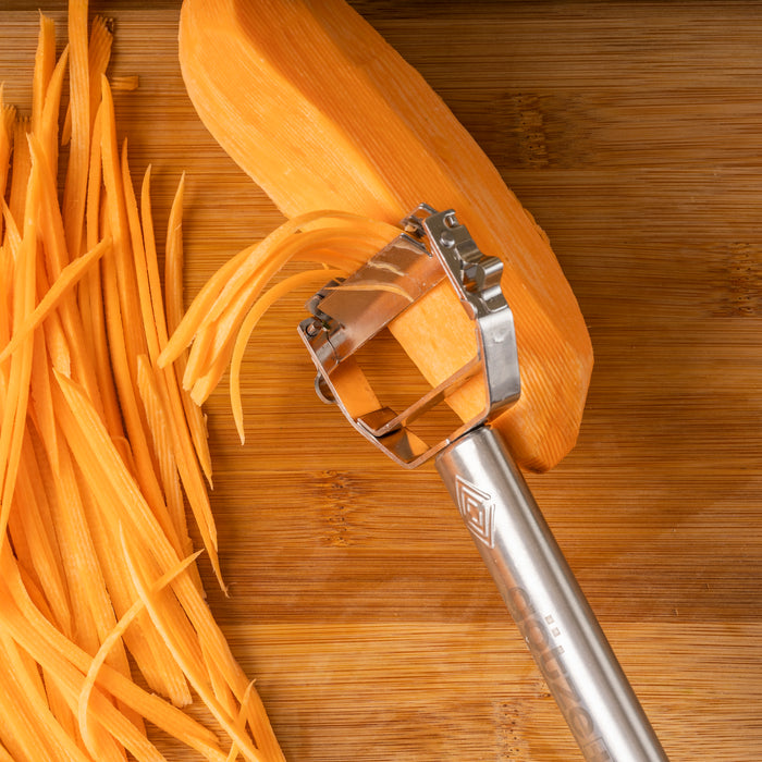 Götze stainless steel julienne peeler creating thin carrot strips on a wooden cutting board, showing the serrated blade in use.