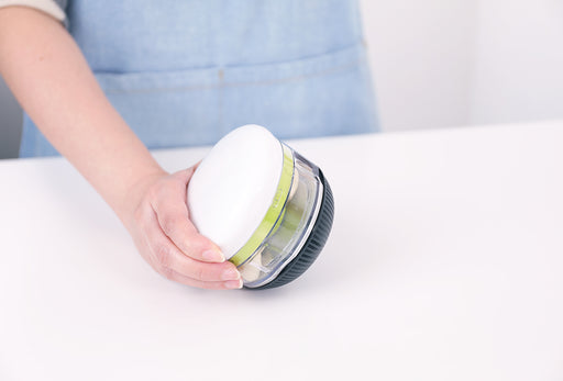 Götze Fresh Mavo manual food chopper held in hand, featuring a clear container, white lid, green blade system, and black non-slip base.