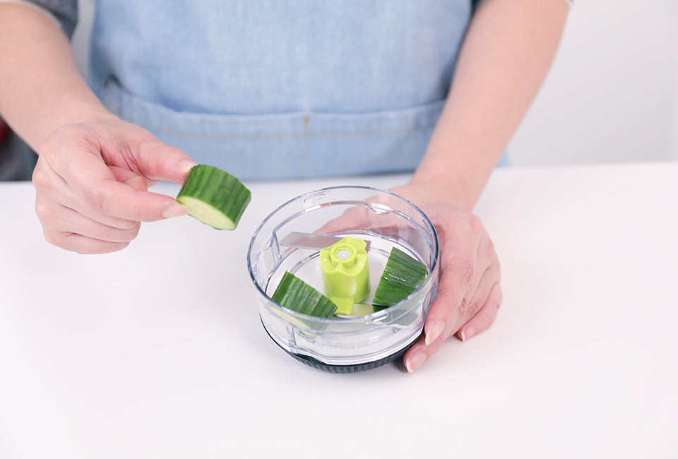 Götze Fresh Mavo manual food chopper with fresh cucumber pieces placed inside the clear container around the green chopping blade.
