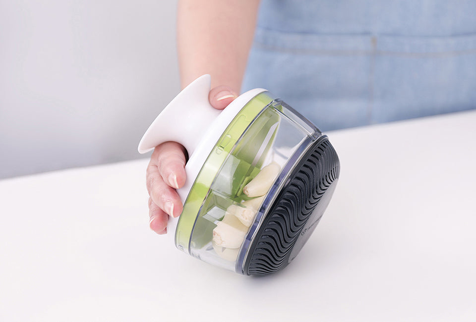 Götze Mavo Sauce Knife held in hand with garlic cloves inside the clear container, featuring green blades and a black non-slip base.