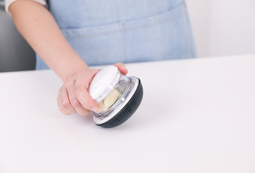 Götze Mini Mavo manual food chopper held in hand with garlic pieces inside the clear container, featuring a white lid and black non-slip base.