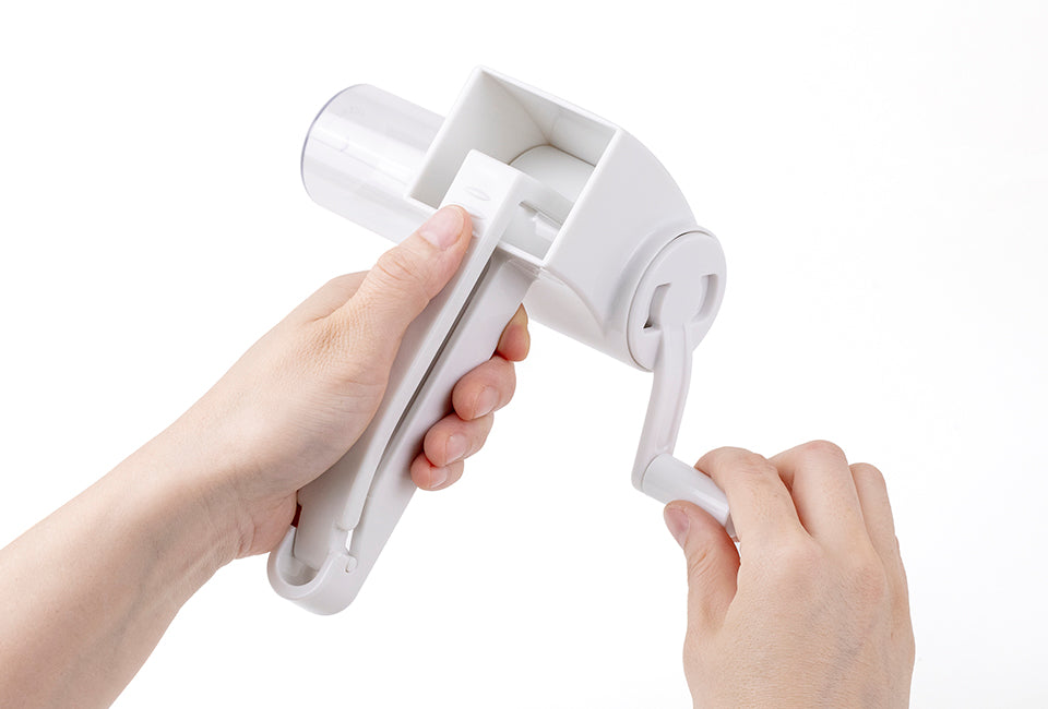 Götze rotary grater held in both hands while turning the hand crank, demonstrating its manual grating mechanism and ergonomic handle design.