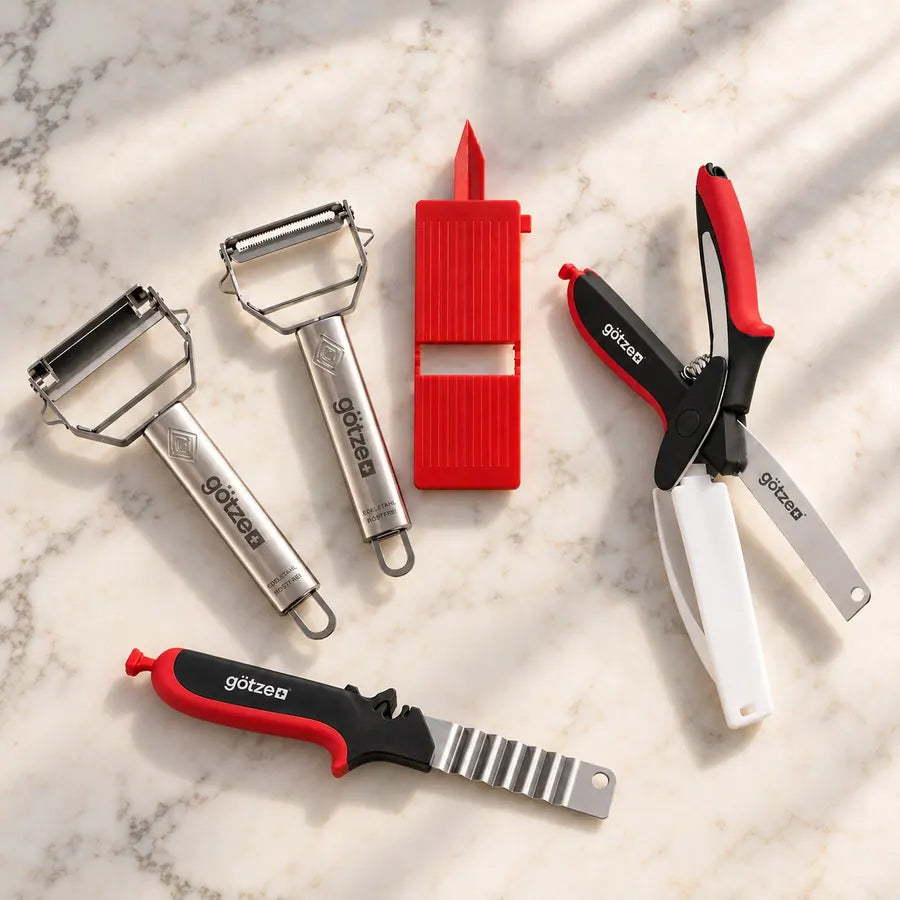 Götze Limited Bundle: two stainless steel peelers, a red mini slicer, 3-in-1 kitchen shears and a crinkle-edge knife on a marble counter
